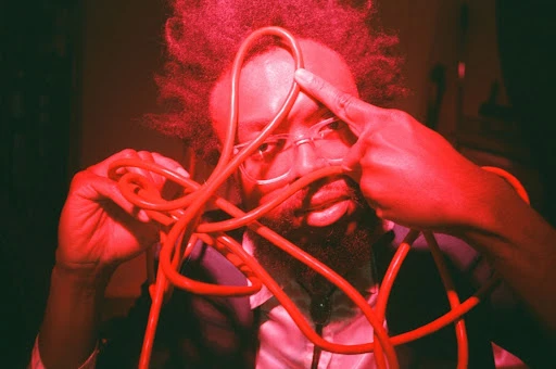 A man with an afro and glasses manipulates a red, coiled cable in a dimly lit indoor setting under bright, saturated red light.