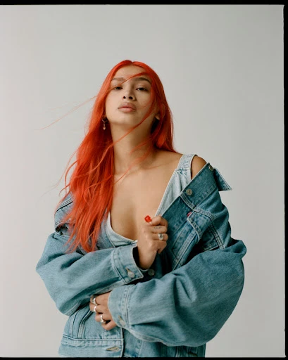 Portrait of a young adult woman with bright red hair wearing a denim jacket against a neutral backdrop in a studio setting.