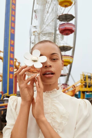 Model with a shaved head wearing a white ruffled blouse holding a flower perfume bottle covering one eye in front of a carnival ride