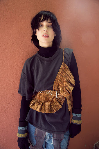 Portrait of a woman with dark hair wearing a grey t-shirt, black long-sleeve shirt, jeans, and a brown velvet ruffled belt and top against a solid wall.
