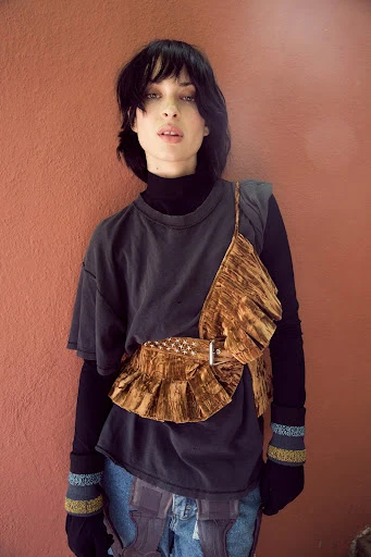 Portrait of a woman with dark hair wearing a grey t-shirt, black long-sleeve shirt, jeans, and a brown velvet ruffled belt and top against a solid wall.