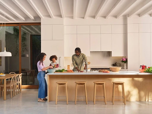 Family of three cooking together in a bright, modern kitchen. An adult female and child stand at a kitchen island while the father chops vegetables.