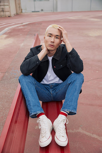 Asian man with blond hair sits on red surface in jeans, black jacket, white shirt, and white sneakers in an outdoor setting.