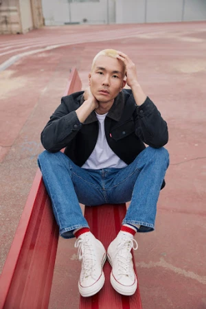 Asian man with blond hair sits on red surface in jeans, black jacket, white shirt, and white sneakers in an outdoor setting.