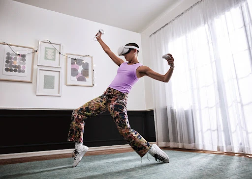 Woman wearing a VR headset and holding controllers, dressed in patterned pants and a tank top, is standing in a brightly lit room.