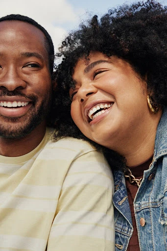 Close-up shot of an African-American couple smiling outdoors; the woman is wearing a denim jacket and gold earrings.