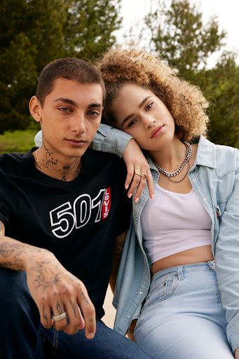 Young couple in casual clothing posing outdoors. Man in 501 tee and jeans, woman in denim jacket and jeans, green trees in background.