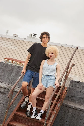 A stylish couple poses on a rooftop staircase with an urban backdrop. The focus is on fashion and an outdoor setting with neutral tones.