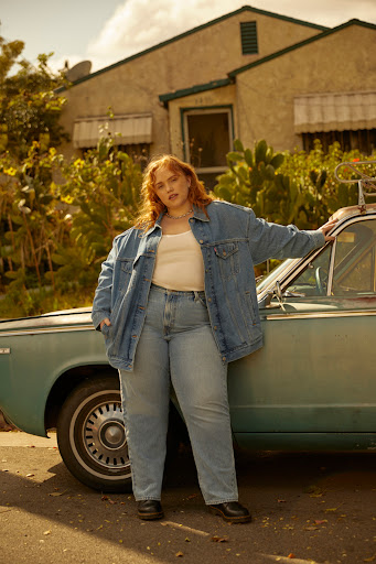 Red-haired woman in a denim jacket and jeans poses next to a vintage car in front of a house on a sunny day. Fashion shot.