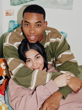 A young couple smiles and embraces warmly in an indoor setting, wearing casual sweatshirts in a close-up shot.