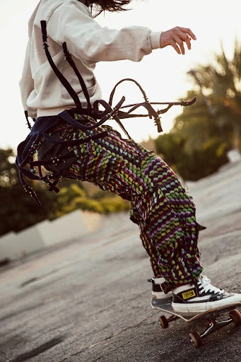 Person in multicolor pants rides skateboard on concrete, with palm trees in the background, wearing a hoodie and sneakers.