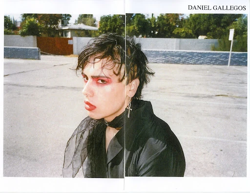 Portrait of a person with dark hair and makeup, wearing a black top and choker, outdoors on an overcast day with buildings in the background.