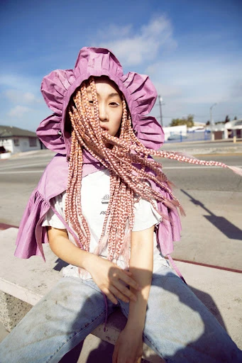 Asian woman sits outside, wearing a pink floral hood and bleached braids. The photo captures an urban street scene with buildings under a blue sky.