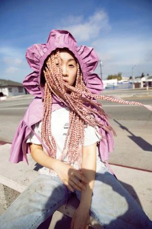 Asian woman sits outside, wearing a pink floral hood and bleached braids. The photo captures an urban street scene with buildings under a blue sky.