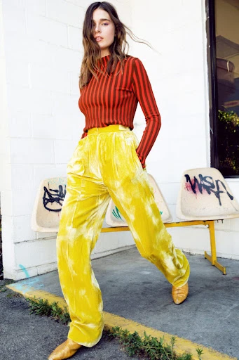 Woman in a red-striped top and yellow velvet pants poses in front of a concrete wall with graffiti and yellow chairs.