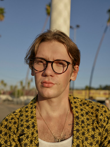 Portrait of a young adult male wearing glasses and a patterned yellow jacket against a backdrop of palm trees and a bright blue sky.