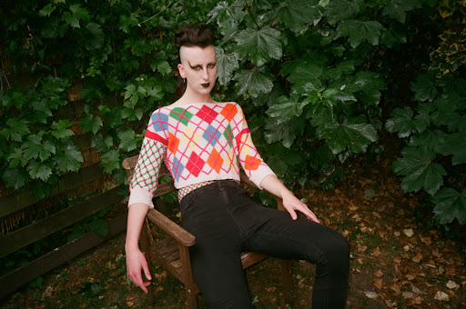 Person with dramatic eye makeup and a patterned sweater poses on a wooden chair against a leafy green backdrop outdoors.