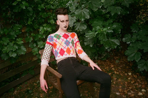 Person with dramatic eye makeup and a patterned sweater poses on a wooden chair against a leafy green backdrop outdoors.