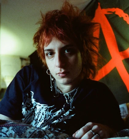 Close-up portrait of a person with spiky red hair and facial piercings wearing a black graphic t-shirt against a dark background.