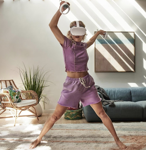 A woman wearing a VR headset and holding controllers stands with legs apart in a brightly lit living room with minimalist decor.