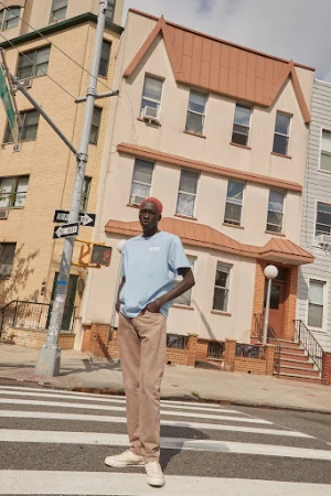 Adult male standing on a crosswalk in an urban environment, wearing a light blue t-shirt, brown pants, and white sneakers.