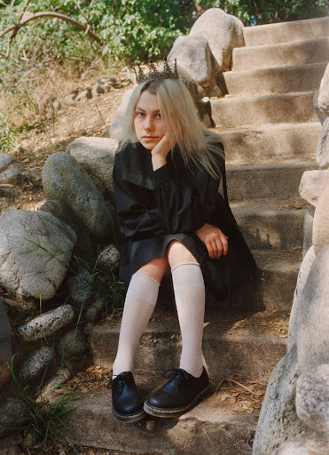A young adult female with platinum blonde hair and a dark crown sits on stone steps surrounded by greenery in a fashion forward editorial shot.
