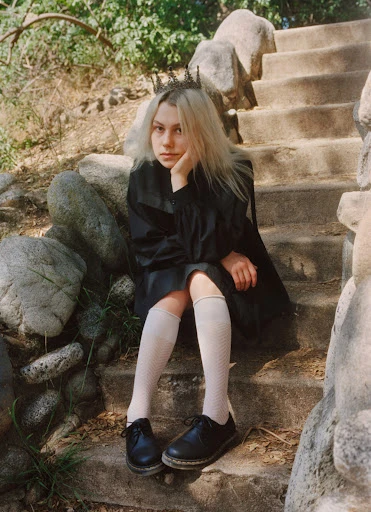 A young adult female with platinum blonde hair and a dark crown sits on stone steps surrounded by greenery in a fashion forward editorial shot.