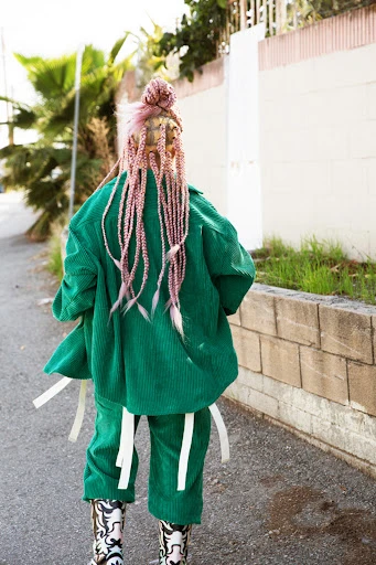Person in green corduroy suit with white-trimmed cowboy boots walks away on paved road next to wall; sunny day, high-angle shot.
