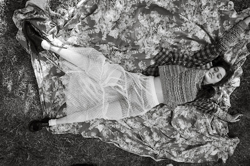 Overhead shot of a woman with dark hair lying on a floral blanket in grass, wearing a knit top and sheer skirt, eyes closed.
