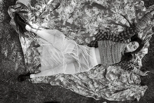 Overhead shot of a woman with dark hair lying on a floral blanket in grass, wearing a knit top and sheer skirt, eyes closed.