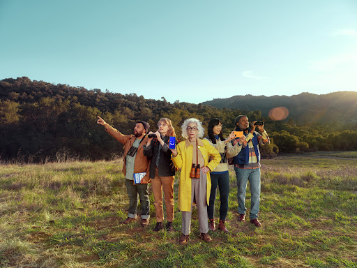 A group of five people of diverse ages and ethnicities stand in a field, surrounded by grassy hills, enjoying a sunny day outdoors.