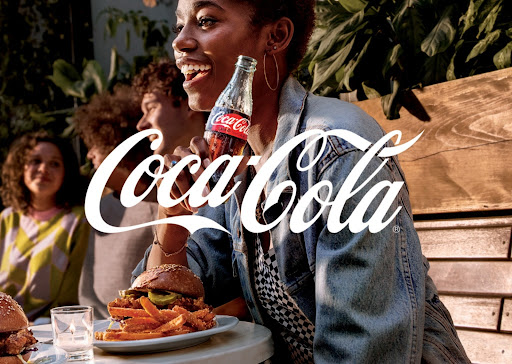 Smiling woman holding a Coca-Cola bottle at an outdoor table with burgers and fries, with other people and plants in the background.