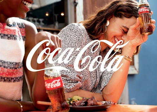 Two cheerful women laugh at an outdoor table while enjoying Coca-Cola and food on a bright day, with branding prominently displayed.