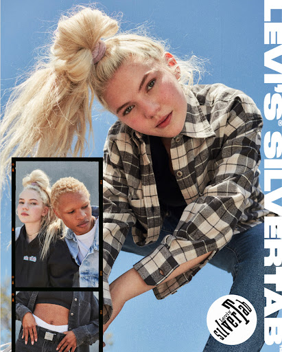 A young woman with blonde hair in a high ponytail poses wearing a plaid shirt and jeans against a sky-blue backdrop in an advertisement.