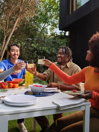 A group of four friends of diverse ethnicities raise glasses at an outdoor table laden with fruit, plates, and beverages.