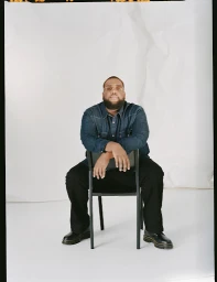 Man with beard wearing denim shirt sitting backwards on a chair against a white backdrop