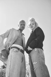 Low-angle monochrome photograph of two models standing outdoors against a bright sky, styled by Gena Tuso.