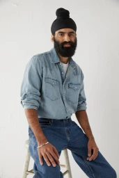A bearded man wearing a black turban, denim shirt, and blue jeans sits on a white stool against a textured white wall.