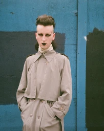 Model with dramatic dark eye makeup and a beige trench coat standing before a blue paneled wall.