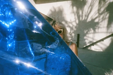 Blonde woman wearing white cat-eye sunglasses behind a translucent blue plastic sheet