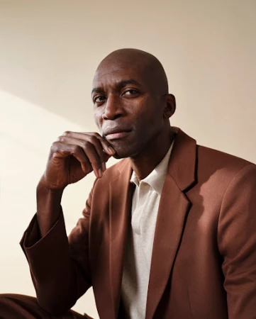 Bald black man in a brown blazer posing with his hand to his chin against a neutral studio backdrop