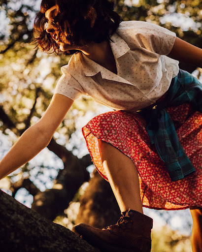 Woman in patterned shirt and red floral skirt climbs a tree in a sunlit outdoor setting, showcasing adventure and outdoor fashion.