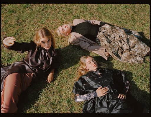 Three young women lie on a grass lawn wearing stylish clothes, captured in a medium shot with soft, natural lighting and a focus on fashion.