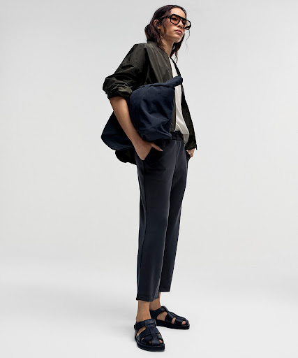 Model wearing dark glasses, olive jacket, and carrying a large navy bag against a white background