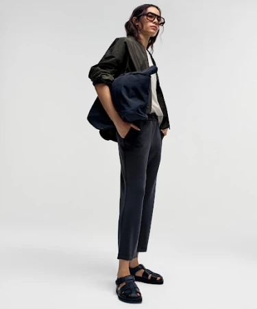 Model wearing dark glasses, olive jacket, and carrying a large navy bag against a white background