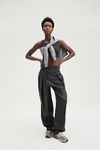 Full-length shot of an adult Black woman wearing a crop top, trousers, sweater around shoulders, and running shoes on a white background.