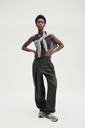 Full-length shot of an adult Black woman wearing a crop top, trousers, sweater around shoulders, and running shoes on a white background.