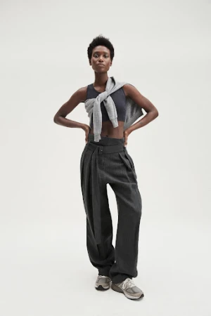 Full-length shot of an adult Black woman wearing a crop top, trousers, sweater around shoulders, and running shoes on a white background.
