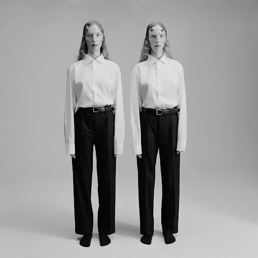 Two people stand side-by-side in a photo studio wearing long-sleeved shirts and dark pants against a neutral grey background in a studio shot.