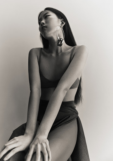 East Asian woman wearing large silver hoop earrings and a dark top seated against a plain light wall.